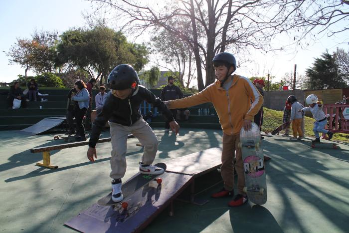 un niño en skate bajando una rampa y otro de pie al lado con el skate en la mano que lo agarra