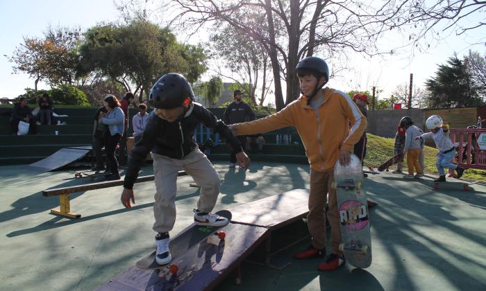 un niño en skate bajando una rampa y otro de pie al lado con el skate en la mano que lo agarra